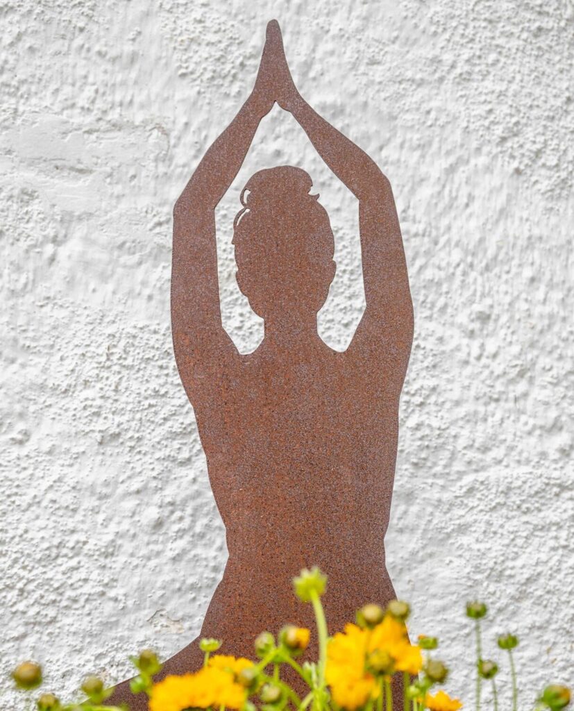 Rostige Metall-Silhouette einer Yoga-Figur zwischen gelben Blumen vor der Hausfassade