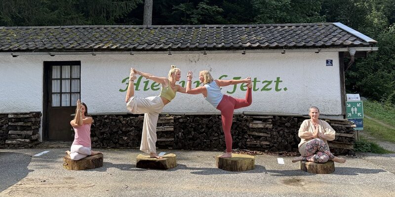 4 Personen machen vor dem Schriftzug „Im Hier und Jetzt“ Yoga